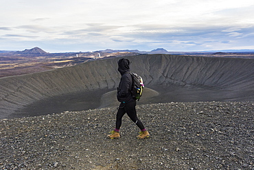 A young woman dressed in black and wearing boots walks along the ridge of the Hverfjall crater with steam rising in the volcanic background.