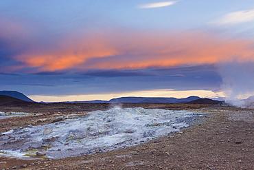 A blue coloured boiling hot mud pool emanates steam as the sunset colours the sky orange, in Hverarâândor Hverir Geothermal Area at Namaskard Pass, North Iceland.