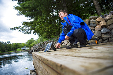 Man crouching on harbor and setting up solar panel to recharge battery, Washego, Ontario, Canada