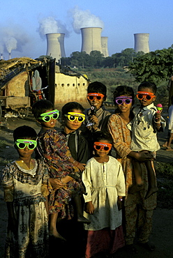 A group of young children gather outside their humble dwellings in India with a Nuclear Plant Facility in the background.