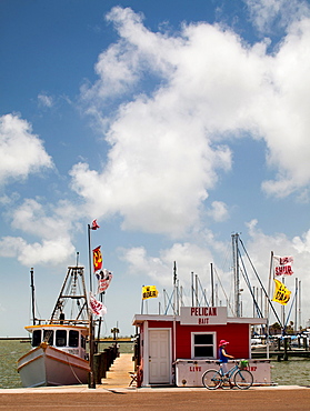 ROCKPORT, TEXAS, USA. A woman wearing a pink floppy hat walks her bike in front of a fishing boat on a sunny day at a harbor.