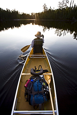 A woman paddles her canoe as the sun rises over the trees on the horizon of the Boundary Waters in Minnesota.