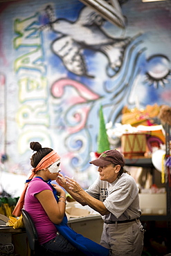 A woman creates a mask on a girl for a parade in Santa Barbara. The parade features extravagant floats and costumes.