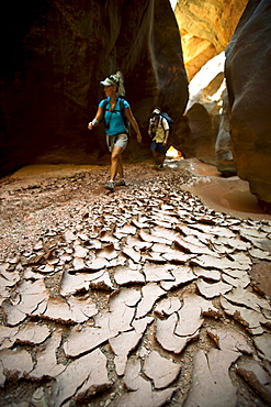 A man and woman hiking through a narrow canyon past dried and cracked mud in Utah.