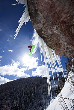 A male snowboarder jumps off an ice waterfall cliff on a sunny day in Colorado.