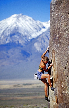 Young woman bouldering in sun with snow capped mountain background.