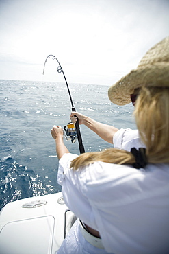 A blonde woman in a straw hat reels in a fish with blue water in the distance.