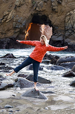 A woman balancing on a rockin front of a blow hole with the setting sun coming through, Pfeiffer Beach, Big Sur, California.