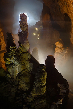 A caver is dwarfed by massive stalagmites in Hang Son Doong. Light can be seen coming in in the distance through the frist doline which is over 3 km away. Hang Son Doong is located in Phong Nha Ke Bang National Park, Vietnam.