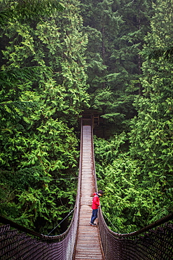 A man in a red coat and jeans stands in middle of a suspension bridge surrounded by large green trees.