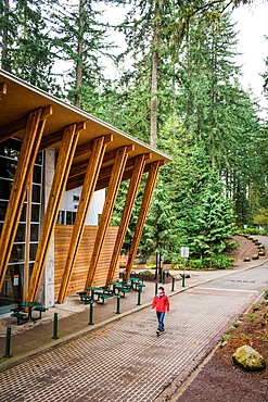 A man in red coat and jeans, walks beside a modern visitors center in a forested park.