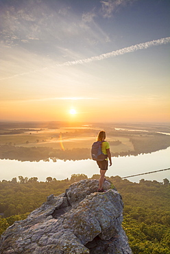 Woman standing on sandstone overhang watching sunrise from summit of Petit Jean Mountain above Arkansas River Valley 