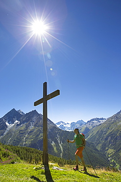 A male hiker is walking towards a wooden cross on a mountain meadow in the Val d'Anniviers in the Swiss Alps. The sun is shining bright, temperatures are high. This is halfway the Haute Route, a classic hike between Chamonix in France and Zermatt in Switzerland. For fit hikers it takes ten days.