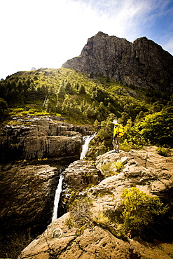 Brody Leven, wearing a vibrant yellow jacket and backpack, overlooks a waterfall flowing through the canyons. Lakes District, Chile.