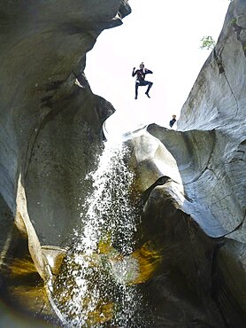 A man in wetsuit jumping from a high cliff next to a waterfall during a canyoning adventure trip in Switzerland. Ticino, in the southern part of Switzerland is a small paradise for canyoning thanks to numerous little waterfalls, torrents and rivers that rush down the valley through the wilderness. Cresciano, Ticino Canton, Switzerland