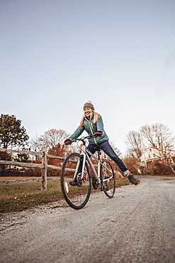 Young woman riding bicycle along countryside dirt road beside wooden fence, Portland, Maine, USA