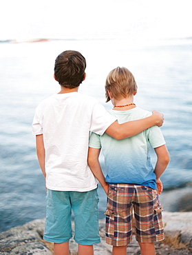 Rear view of two brothers wearing t-shirts and shorts with arm around standing against sea