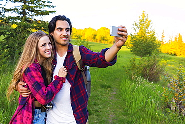 Young couple in a park posing for a self-portrait with their cell phone at sunset; Edmonton, Alberta, Canada