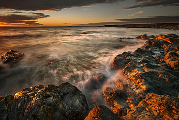 Atlantic ocean water rolls into the rocky shoreline at sunrise with a view of the coastline; Bonavista, Newfoundland, Canada