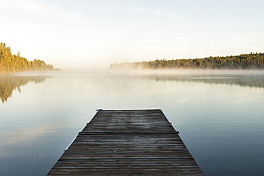 A Wooden Dock Leading Out To A Misty Tranquil Lake In Riding Mountain National Park, Wasagaming, Manitoba, Canada