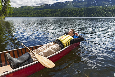 Man Reclined In Canoe And Resting In The Sunshine On Byers Lake With Forested Foothills In The Background, Byers Lake Campground, Denali State Park, Alaska, United States Of America