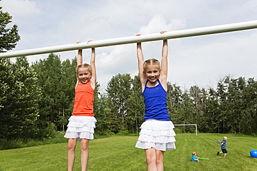 Two Girls Holding Onto The Crossbar Of A Soccer Goal Post In A Park, Stony Plain, Alberta, Canada