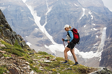 Female Hiker With Poles Hiking Up Mountain Path With Mountain Cliff With Snow In Background, British Columbia, Canada