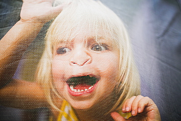 A Young Girl With A Silly Expression On Her Face Viewed Through A Window Screen, Peachland, British Columbia, Canada