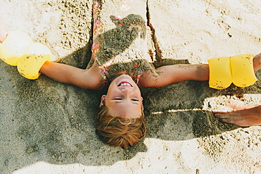 A Young Girl Being Buried In The Sand On The Beach, Peachland, British Columbia, Canada