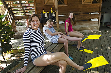 A Mother And Two Daughters Sit On A Wooden Bench With Flippers On Their Feet, Utila Island, Honduras