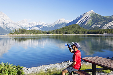 A Man Drinks From A Water Bottle On The Edge Of A Lake Overlooking The Rocky Mountains, Kananaskis, Alberta, Canada