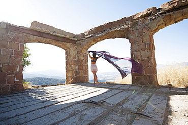 A Woman Holding A Piece Of Sheer Fabric Behind Her As It Flows In The Wind Within Some Ruined Arches, Santa Barbara, California, United States Of America