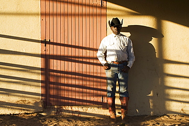 Agriculture - A young black cowboy stands against a bunkhouse wall with late afternoon shadows / Childress, Texas, USA.