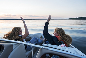 Two Teenage Girls Riding In The Front Of A Boat On A Tranquil Lake At Sunset Giving Each Other A High Five, Ontario, Canada