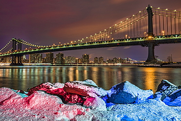 Manhattan Bridge By Snow-Covered Rocks At Sunset, Brooklyn Bridge Park, Brooklyn, New York, United States Of America