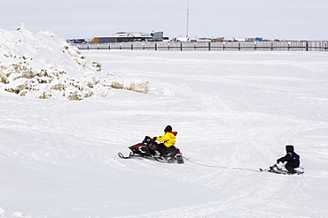 Native Alaskan Youth On A Snowmachine Towing Another On Sled, City Of Barrow In The Background, North Slope, Arctic Alaska, USA, Winter