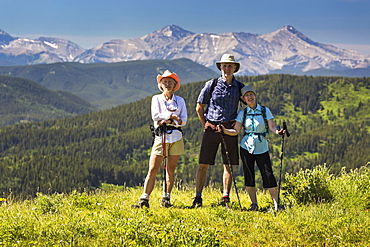 Three Hikers, Two Female And One Male, On Top Of Grassy Hill With Rolling Foothills And Mountain Range In The Background With Blue Sky, Kananaskis Country, Alberta, Canada