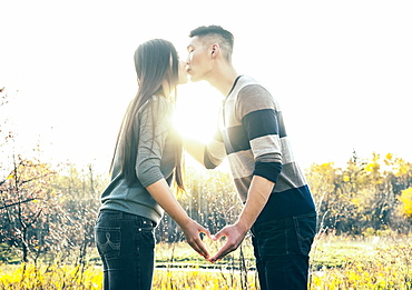 A Young Asian Couple Kissing In A Park In Autumn And Making A Heart With Their Hands In The Warmth Of The Setting Sun, Edmonton, Alberta, Canada