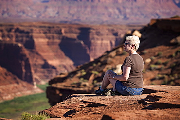 Woman Sitting On The Edge Of A Rock Canyon Cliff Looking Over The Colorado River With Canyon Walls In The Background, Colorado, United States Of America