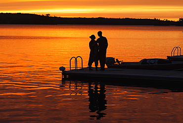 Silhouette Of A Couple Standing At The End Of A Dock At Sunset With An Orange Glow In The Sky And Reflected In The Tranquil Lake Water, Ontario, Canada