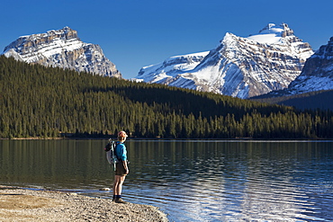 Female Hiker Standing Along A Rocky Shoreline Of A Mountain Lake With Snow Peaked Mountains Reflecting In Lake And Blue Sky, Banff National Park, Alberta, Canada