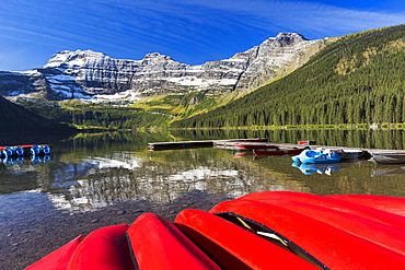 Mountain Lake With Dock Reflecting Snow Peaks And Blue Sky With Upside Down Red Canoes In The Foreground, Waterton, Alberta, Canada