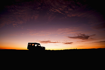 A Truck Parked On The Savannah With It's Headlights On Is Shown In Silhouette On The Horizon At Sunrise, Blue And Orange Sky With A Few Thin Clouds In The Background, Narok, Kenya