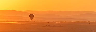 A Hot Air Balloon Flies Over The African Savannah In The Orange Light Before Sunrise, With A Trail Of Dust Below Made By A Truck On The Ground, Narok, Kenya