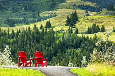 Two Red Adirondack Chairs On A Paved Pathway On Top Of A Hillside Overlooking Rolling Treed Hillside In The Distance, South Of Maple Creek, Alberta, Canada