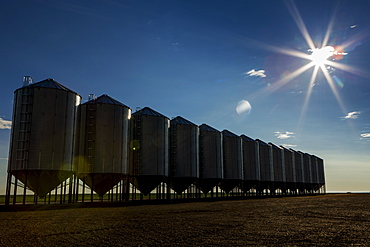 A Row Of Large Metal Grain Bins With A Sun Burst And Blue Sky, Mossleigh, Alberta, Canada