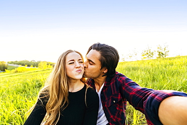 A young couple sits in a field taking a self-portrait while the young man kisses the young woman on the cheek, Edmonton, Alberta, Canada