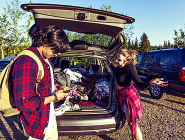 Young couple standing at the back of their packed vehicle with back door open and the young man in using his cell phone while the young woman waits impatiently, Edmonton, Alberta, Canada