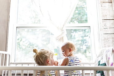 Two sisters, a baby and a toddler, play together in a crib by a window, Sorrento, British Columbia, Canada