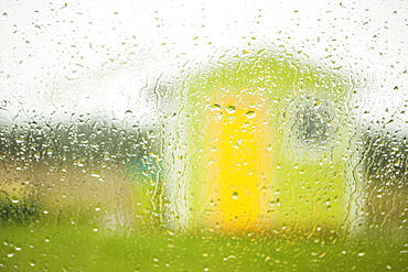 A small green building with yellow door and a window viewed through a window wet with raindrops, Newfoundland, Canada